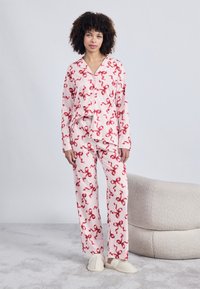 Pijamas cor-de-rosa com padrão de fita vermelha, apresentando uma camisa de botões e calças longas, feitas de tecido suave, combinadas com pantufas bege.