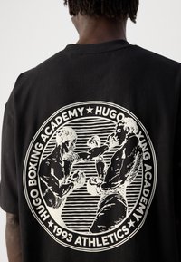 Asmuo su juoda maike, ant kurios nugaros yra baltas apvalus logotipas, vaizduojantis du bokso kovotojus pasiruošusius mūšiui, ir tekstas "Hugo bokso akademija 1993 sportas."