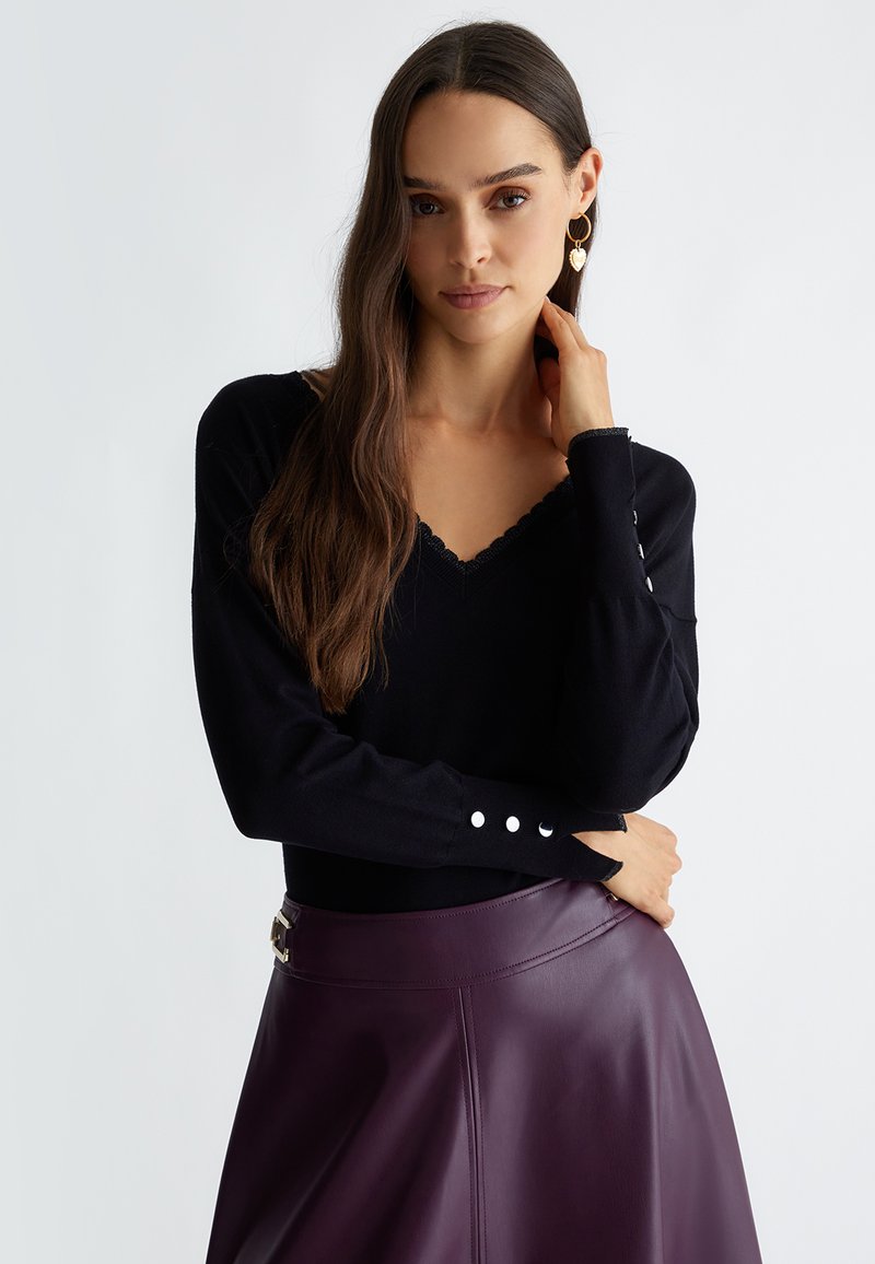 Suéter negro de manga larga con escote en V y ribete con ondas, con detalles de botones en los puños, combinado con una falda de cuero morada oscura.