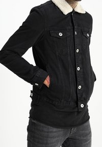 Černá džínová bunda s bílým kožíškovým límcem, zavíráním na knoflíky vpředu, dvěma náprsními kapsami a bočními rozparky pro ruce.