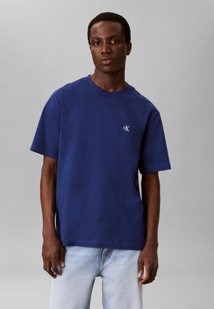 Giovane uomo che indossa una t-shirt semplice blu scuro a maniche corte con un piccolo logo bianco "cK" sul petto e jeans azzurri, in piedi davanti a uno sfondo grigio.
