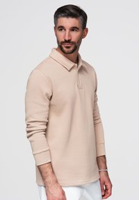 Hombre con el pelo corto y oscuro y barba, lleva un polo beige de manga larga y pantalones blancos, de pie y mirando hacia un lado.