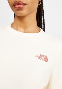 Gros plan sur une personne portant un sweat-shirt crème avec le logo "The North Face" en rose sur le côté gauche de la poitrine et une petite boucle d'oreille en or.