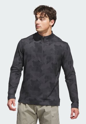 Joven con sudadera de manga larga y media cremallera de camuflaje gris oscuro y pantalones beige claro, mirando hacia su izquierda.