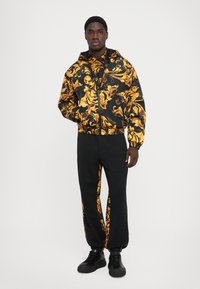 Man som står iklädd en svart och guldmönstrad jacka och matchande svarta byxor med paneler samt svarta sneakers mot en vit bakgrund.