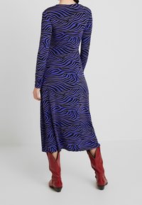 Pitkähihaiset mekko violetissa ja mustassa seepra-kuosissa, valmistettu joustavasta kankaasta. Yhdistetty polven korkeisiin punaisiin saappaisiin, jotka korostavat virtaavaa siluettia.