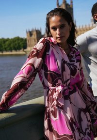 Šaty s dlouhým rukávem a knoflíkovou přední částí v růžovém a hnědém květinovém vzoru. Obsahují stažený pas s páskem a lehkou, splývavou tkaninu.