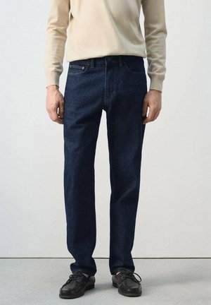 Mand iført mørkeblå jeans, beige langærmet skjorte og sorte lædersko, stående mod en ensartet lys baggrund.