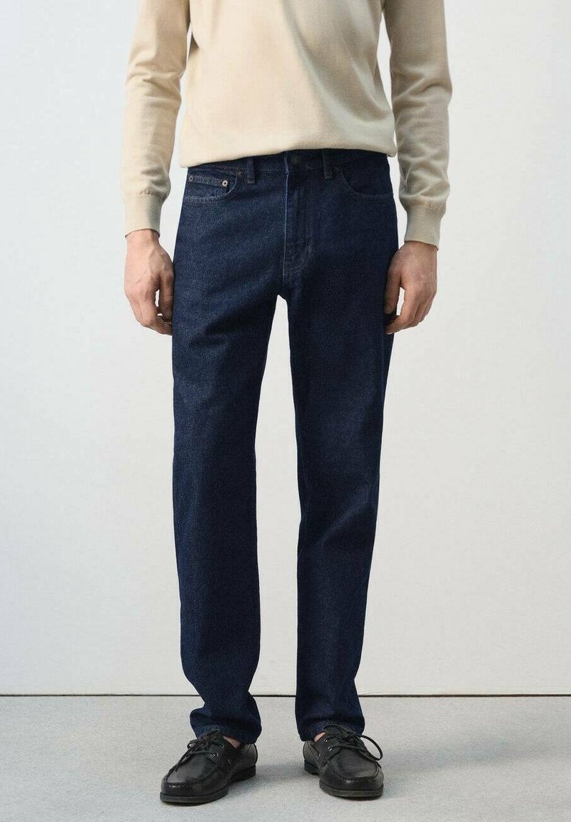 Homme portant un jean bleu foncé, une chemise beige à manches longues et des chaussures en cuir noires, debout devant un fond clair uni.