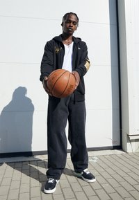 Sudadera negra con cremallera y un logo, camiseta blanca, pantalones oscuros holgados y zapatillas negras, sosteniendo un balón de baloncesto marrón al aire libre sobre una superficie pavimentada.