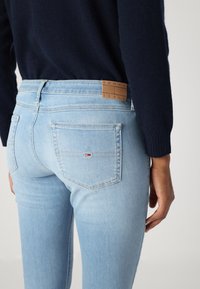 Világoskék farmernadrág slim fit szabással. Két hátsó zsebbel és "TOMMY JEANS" felirattal ellátott barna bőrcímkével rendelkezik. Sima textúrájú.