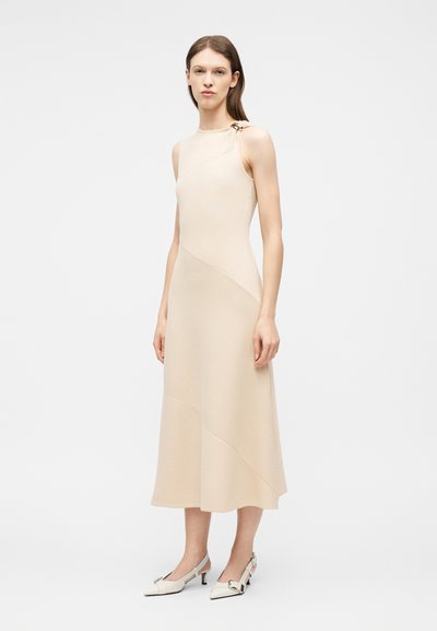 Femme portant une robe midi beige sans manches avec une texture côtelée asymétrique et un détail anneau doré sur une épaule, associée à des chaussures blanches à talons.