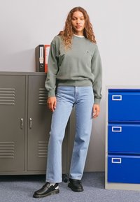 Green sweatshirt, light blue straight-leg jeans, and black slip-on shoes. Background features gray lockers and a blue filing cabinet.