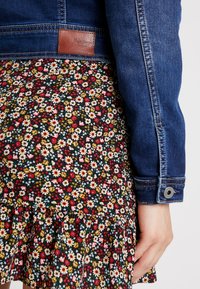 Jeansjacka med en mörkblå finish, med en brun läderetikett; lagd över en blommönstrad svart klänning med volangkant.