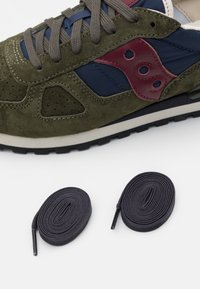 Olivgrön mockaskor med marinblå detaljer och bordeauxfärgade logotypdetaljer. Inkluderar två rullade svarta skosnören på en vit bakgrund.