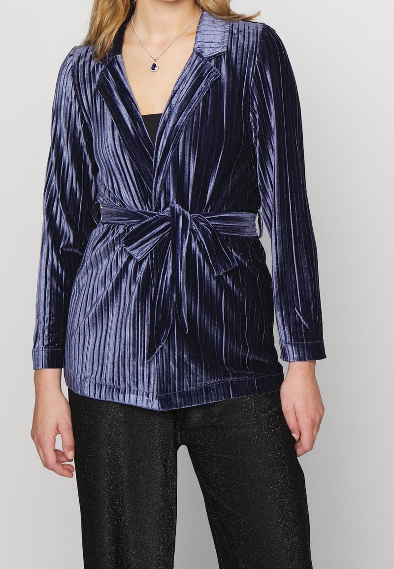 Navyblauwe fluwelen blazer met verticale ribbelstructuur, met notch revers en een strikceintuur op de taille. Lange mouwen; gecombineerd met zwarte broeken.