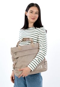 Beige tekstuuri käsilaukku, jossa on kaksinkertaiset kahvat, etutasku vetoketjulla ja säädettävät sivuhihnat, esitetty valkoista taustaa vasten.