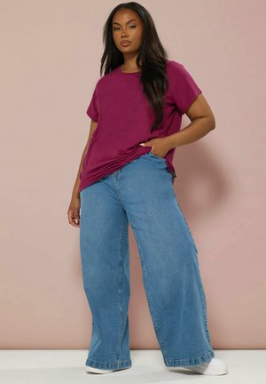 Mujer con una camiseta holgada de color magenta, vaqueros de pierna ancha azul y zapatillas blancas, de pie con una mano en el bolsillo contra una pared beige.