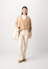 Beige strikkegenser, hvit skjorte med krage, kremfargede bukser og brune slip-on sko. Modellen holder en lys beige skulderveske.