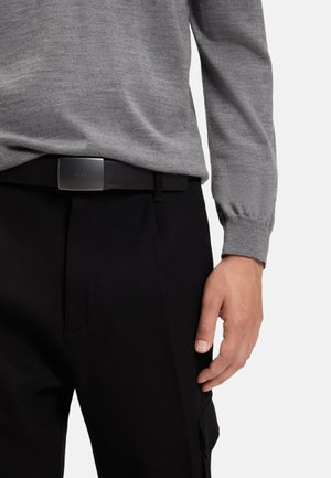 Suéter gris de punto con mangas largas, combinado con pantalones negros y un cinturón de cuero negro con una hebilla metálica rectangular con el logotipo.