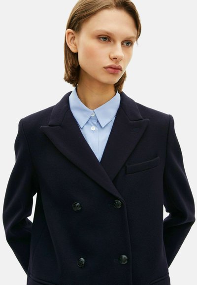 Donkerblauwe dubbelgebreide jas met een scherpe kraag, voorzien van zwarte knopen en een borstzak, gedragen over een lichtblauw overhemd met kraag.