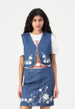 Denim vest with rounded edges and floral embroidery, paired with a matching denim skirt featuring similar floral patterns. White T-shirt underneath.