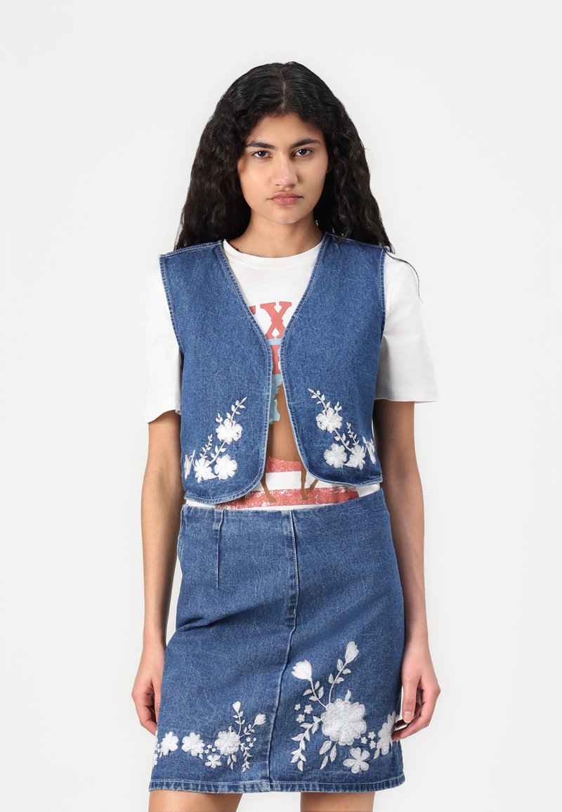 Colete de denim com bordas arredondadas e bordados florais, combinado com uma saia de denim correspondente que apresenta padrões florais semelhantes. T-shirt branca por baixo.