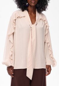 Jasny beżowy bluzka z falbanami przy rękawach i dekolcie. Miękka, lekka tkanina z wiązanym przodem. Połączona z ciemnobrązowymi spodniami.