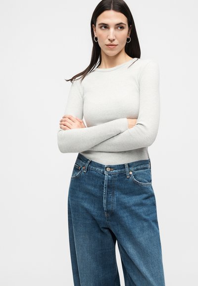 Γυναίκα με σκούρα μαλλιά που φοράει ασημένια κρίκους σκουλαρίκια, ανοιχτό γκρι ριπ τοπ με μακριά μανίκια και φαρδιά μπλε τζιν, στέκεται με σταυρωμένα τα χέρια.