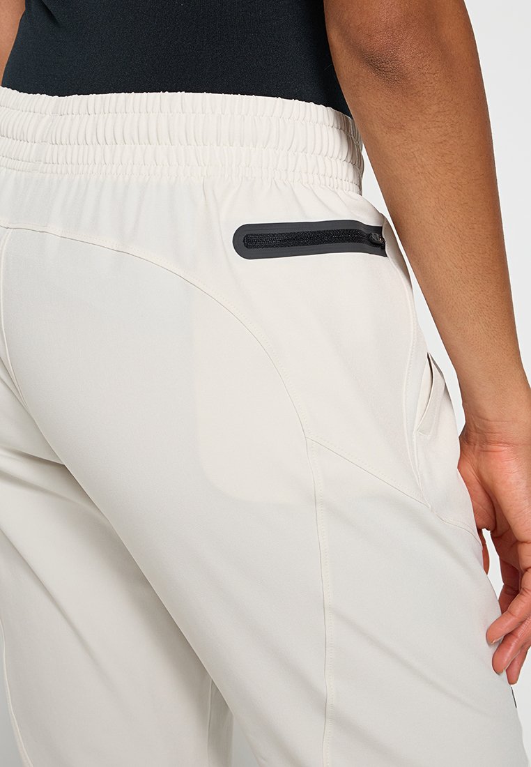 Primo piano di una persona che indossa pantaloni beige chiaro con una cintura elastica e una tasca con zip nera sul lato destro posteriore.