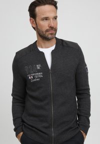 Pilkas megztinis su užtrauktuku, su juodais odos peties akcentais ir siuvinėtu tekstu "924 REG. TM" bei Prancūzijos vėliava.