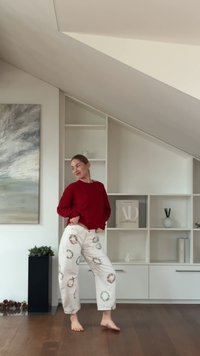 Pulover roșu cu mâneci lungi asortat cu pantaloni crem cu modele florale circulare. Podea din lemn și unitate de rafturi albe în fundal.