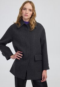 Cappotto di lana grigio scuro con colletto a punta, tasche frontali e maniche larghe, leggermente accorciate. Tessuto liscio e vestibilità su misura.