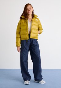 Rumena napihnjena jakna z visokim ovratnikom, prikazana nad belo rebrasto majico, v kombinaciji z mornarsko modrimi cargo hlačami in metalnimi supergami.