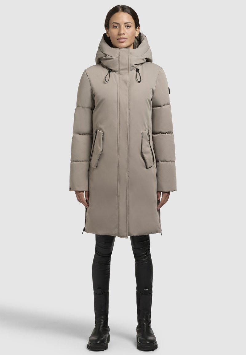 Beige gepolsterter Mantel mit Kapuze, ausgestattet mit einem Frontreißverschluss, Seitentaschen und Kordeln. Trägt er über schwarzen, figurbetonten Hosen und Stiefeln.