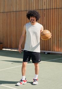 Sportos férfi világosszürke ujjatlan pólóban, fekete logós nadrágban, fehér zokniban és piros részletekkel ellátott sportcipőben, kezében kosárlabdával.