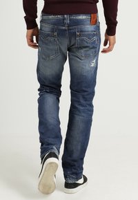 Vaqueros de mezclilla azul oscuro con un acabado desgastado, que presentan bolsillos traseros y un sutil desgaste en las piernas. Llevados con zapatillas de deporte negras y blancas.