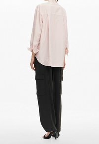 Hellrosa gestreifte Bluse mit lockerem Schnitt, rundem Saum und hochgekrempelten Ärmeln, kombiniert mit schwarzen, weit geschnittenen Cargohosen und Sandalen mit hohen Absätzen.