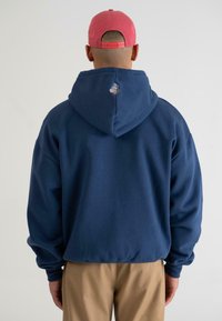 Felpa blu navy con un ampio cappuccio, polsini a coste e un design multicolore ricamato sul retro. Abbinata a pantaloni khaki.
