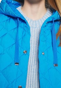 Närbild av en person som bär en ljusblå stickad tröja under en ljusblå vadderad jacka med huva och dragsnören.