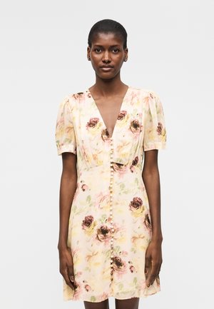 Nő krémszínű, rövid ujjú ruhát visel, amely rózsaszín és sárga virágmintával, valamint gyöngygombokkal díszített az elején.