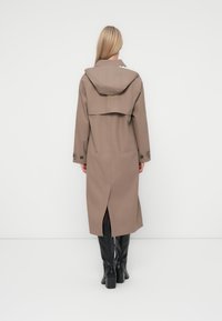 Long, hooded trench coat in taupe, featuring a double-breasted design, side slits, and button accents at the cuffs. Paired with black knee-high boots.