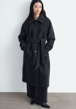 Femme aux longs cheveux noirs portant un manteau en laine gris foncé ceinturé sur un large pantalon noir et des chaussures noires pointues, debout contre un mur blanc.