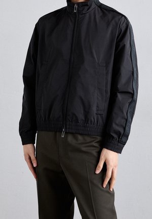 Mężczyzna noszący czarną kurtkę na zamek błyskawiczny z elastycznymi mankietami i dołem, z markowymi paskami na rękawach, w połączeniu z ciemnymi spodniami.