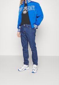 Modrá prošívaná bunda s bílými pruhy, černé tričko s logem, modré džínové kalhoty a bílé tenisky se šedými detaily.