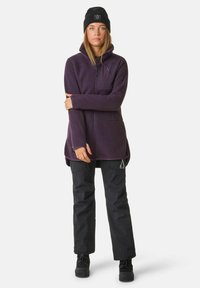 Fleecejacke in Dunkelpurpur mit Reißverschluss vorne, hohem Kragen und Brusttasche. Kombiniert mit schwarzen Hosen und einer schwarzen Mütze. Strukturierter Stoff.