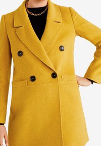 Mosterdgele double-breasted jas van zacht wol, met zwarte knopen, ingezette revers en zijzakken. Soepele, gestructureerde uitstraling.