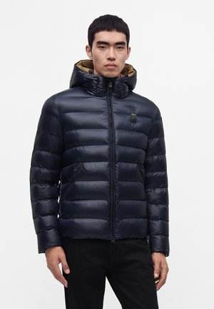 Jonge man die een donkerblauwe gewatteerde jas met capuchon draagt, staande tegen een effen lichte achtergrond, voor zich uit kijkend.