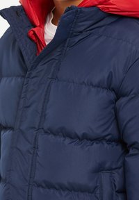 Personne portant une veste matelassée bleu marine avec une couche à capuche rouge visible et une chemise blanche en dessous.