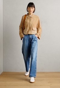 Beige langermet skjorte med lommer, løse blå denimbukser og hvite sko mot en lys bakgrunn og tregulv.